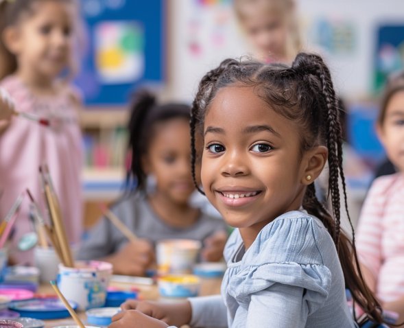 A child smiling in an art classroom with other children blurred out in the background